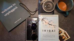 A flat lay of relaxing getaway essentials on a dark table, including a "welcome to unplugged" cabin manual, an instant camera, a mug of coffee, the book "Ikigai", a smartphone with sunglasses, and two small instant photos.