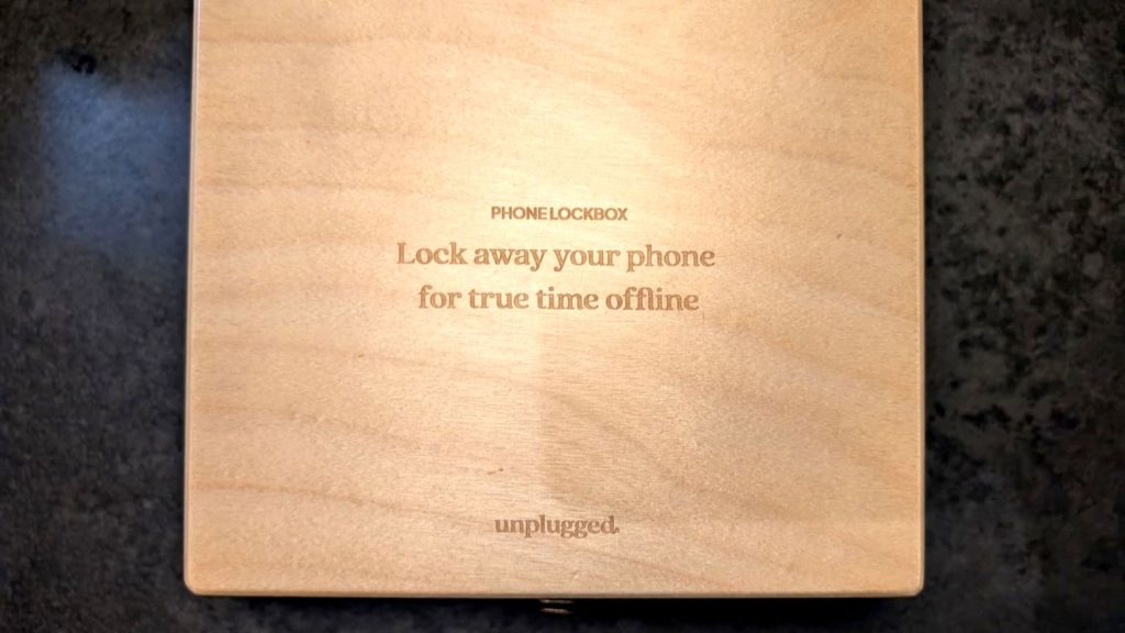 A top-down view of a light wooden box resting on a dark surface. Engraved text on the lid reads "PHONE LOCKBOX. Lock away your phone for true time offline," with the brand name "unplugged." at the bottom