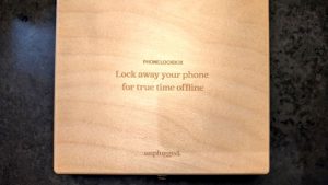 A top-down view of a light wooden box resting on a dark surface. Engraved text on the lid reads "PHONE LOCKBOX. Lock away your phone for true time offline," with the brand name "unplugged." at the bottom