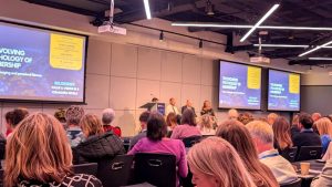 Audience members seated at round tables watching a panel discussion on stage, with presentation screens reading "The Evolving Psychology of Membership" and "Belonging, Value & Vision in a Changing World."
